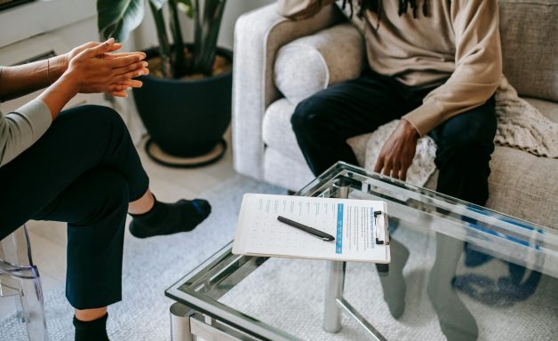 A table with a note pad and pen on it with two people talking in a therapy session