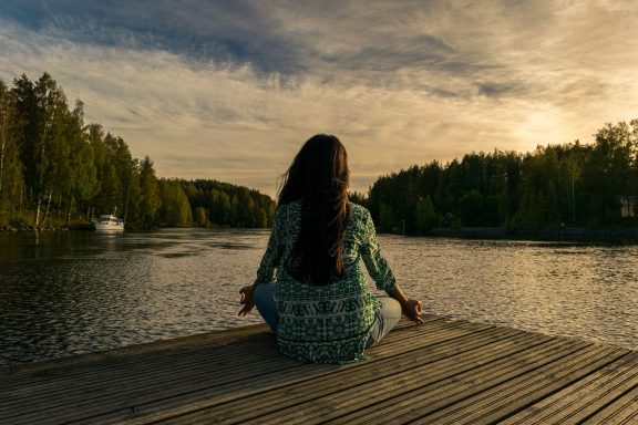 a lady sitting crossed legged next to a lake practicing mindfulness